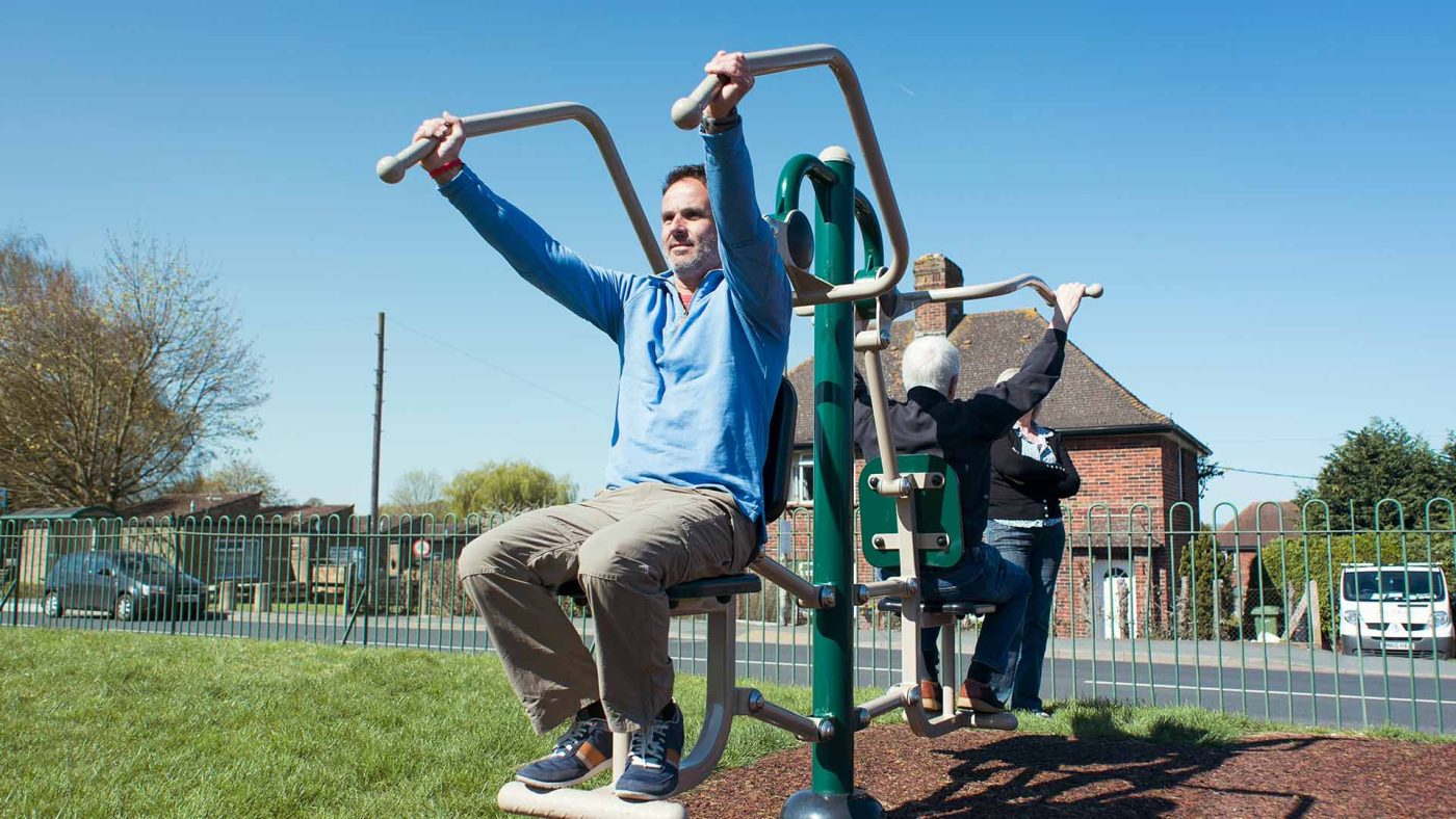 Adults using outdoor fitness equipment on rubber surfacing beside a school