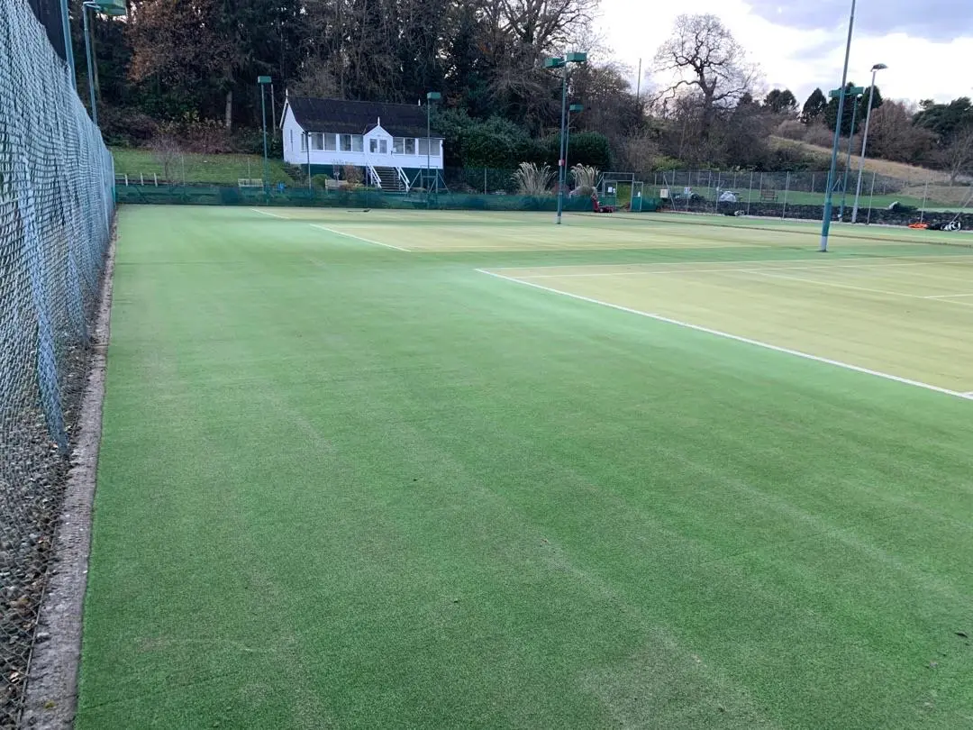 Tennis courts with green turf stretch into the distance. A quaint white clubhouse sits on a hill surrounded by trees, under a partly cloudy sky.