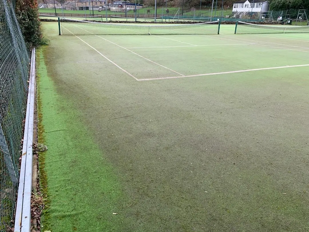 A weathered outdoor tennis court with faded green lines, bordered by a metal fence. The court surface appears worn, creating a nostalgic atmosphere.