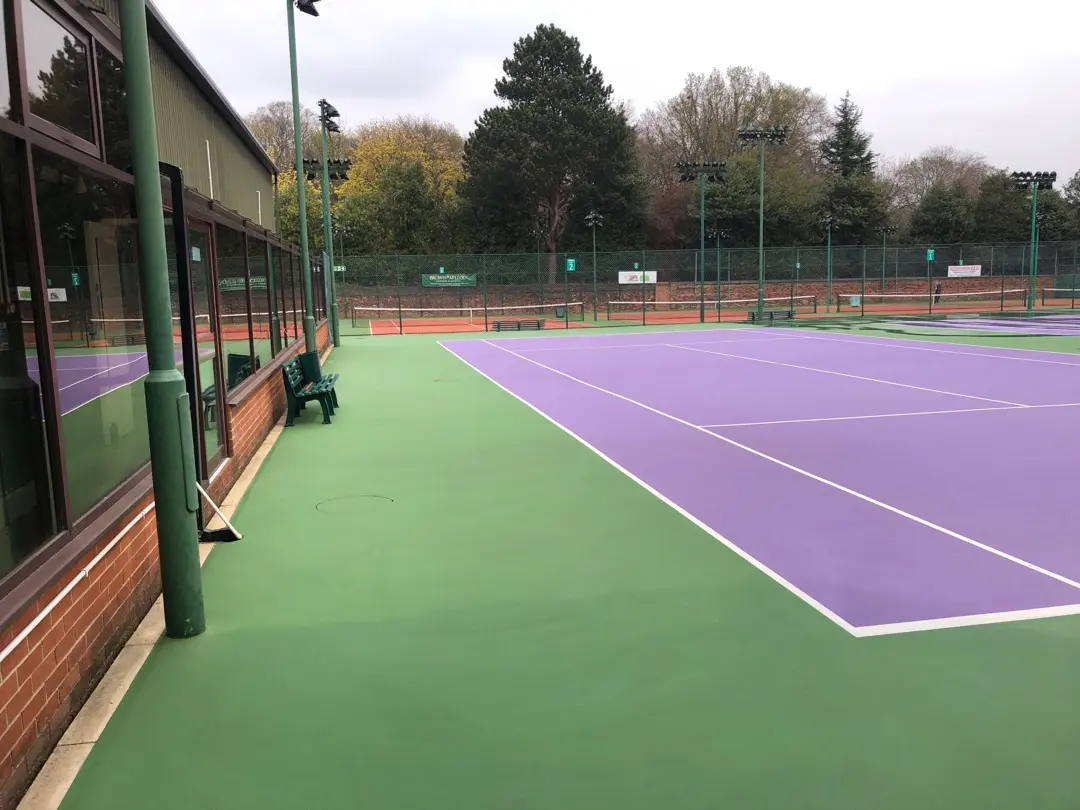 Outdoor tennis court with a vibrant purple playing surface and green borders. Overcast sky and surrounding trees give a calm, serene atmosphere.