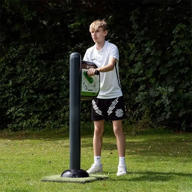 A young person in sportswear uses an outdoor sanitizing station in a park. They stand on grass, with leafy trees in the background, conveying a sunny, active mood.