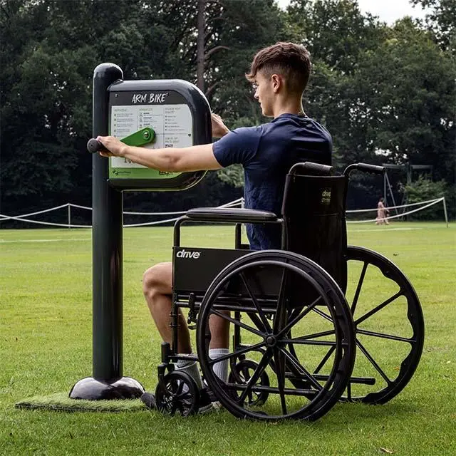 A person in a wheelchair exercises on an outdoor arm bike in a grassy park, exuding determination. Trees and a tennis net are visible in the background.