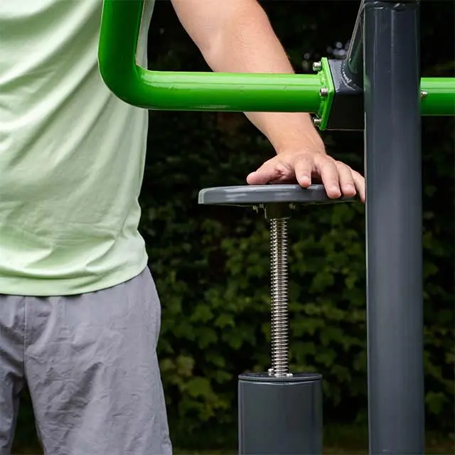 A person in a light green shirt touches a spring-mounted, black outdoor exercise machine. The green metal bars stand out against a leafy background.
