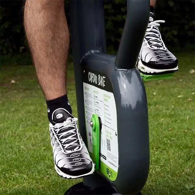 A person wearing black and white athletic shoes is pedaling a stationary "Cardio Bike" in a grassy outdoor setting, conveying a sense of fitness.