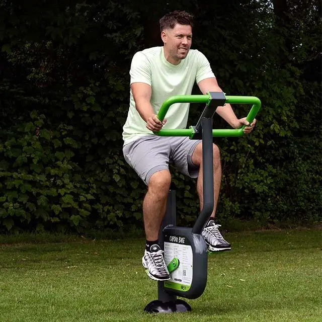 A man on an outdoor exercise bike in a park, wearing a light green shirt and gray shorts. The scene is set against lush green trees, conveying a calm, active vibe.