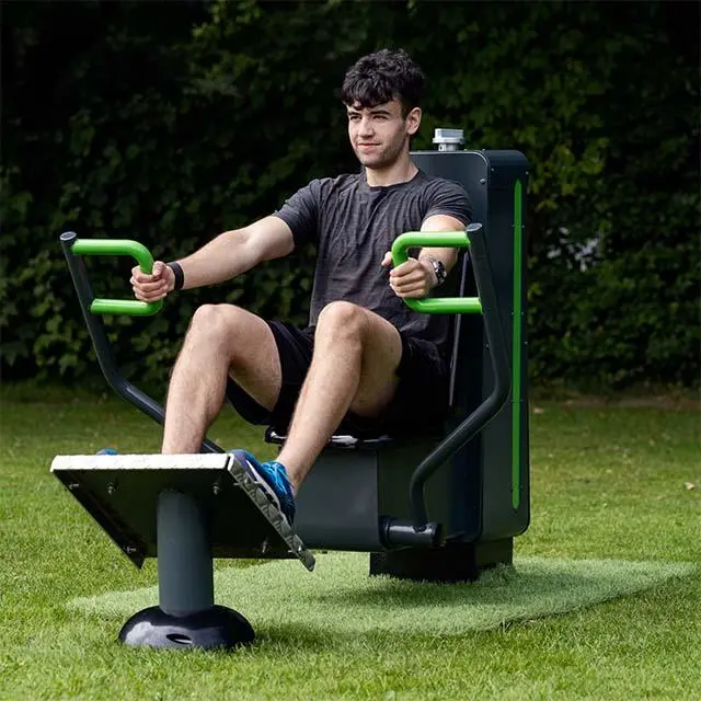 A man exercises on a row machine outdoors. He wears a grey T-shirt and black shorts, with a focused expression. Green grass and bushes are in the background.