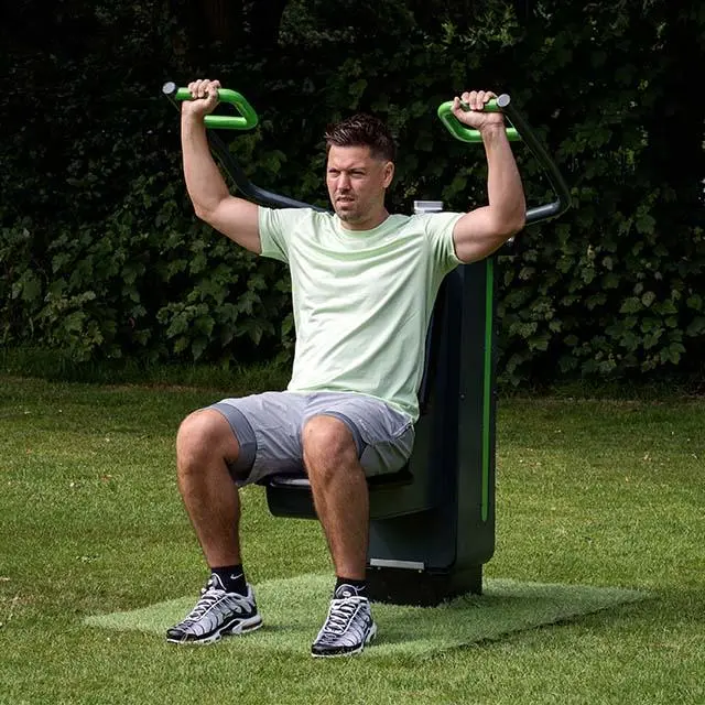 A man exercises on an outdoor fitness machine in a park, wearing a light green T-shirt and gray shorts. He has a focused expression amidst lush greenery.