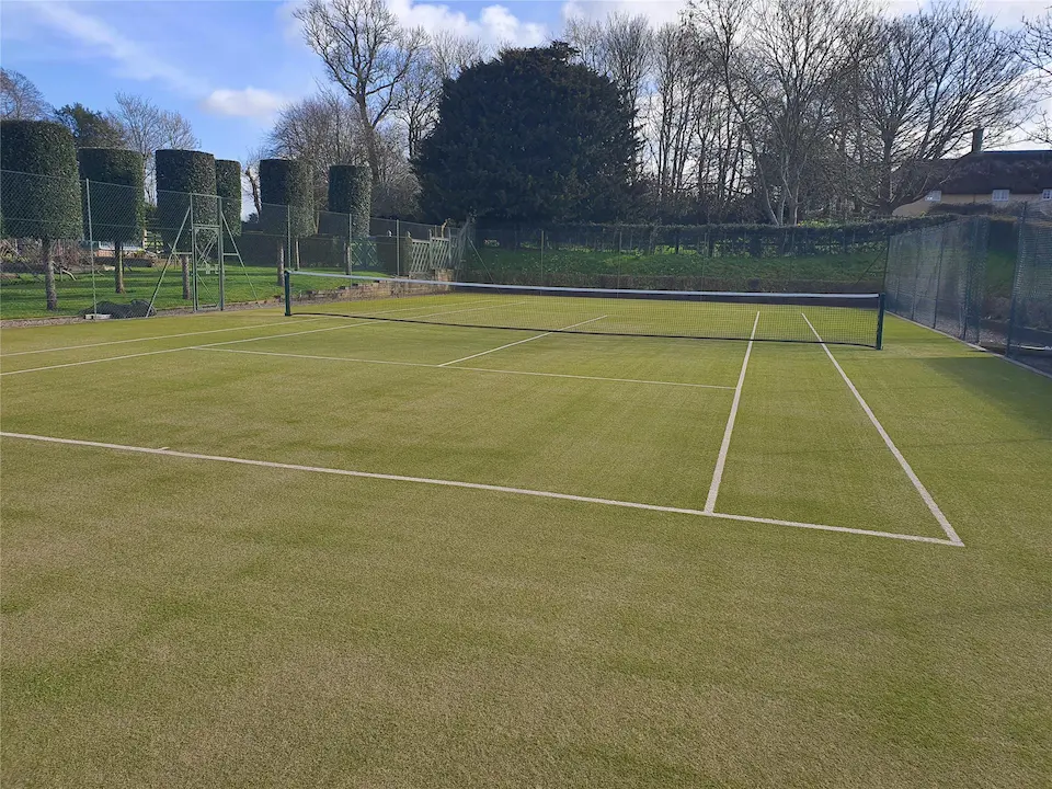 A well-maintained grass tennis court with a net and bright lines, surrounded by a fence and tall trees under a blue sky, conveying a calm, serene atmosphere.