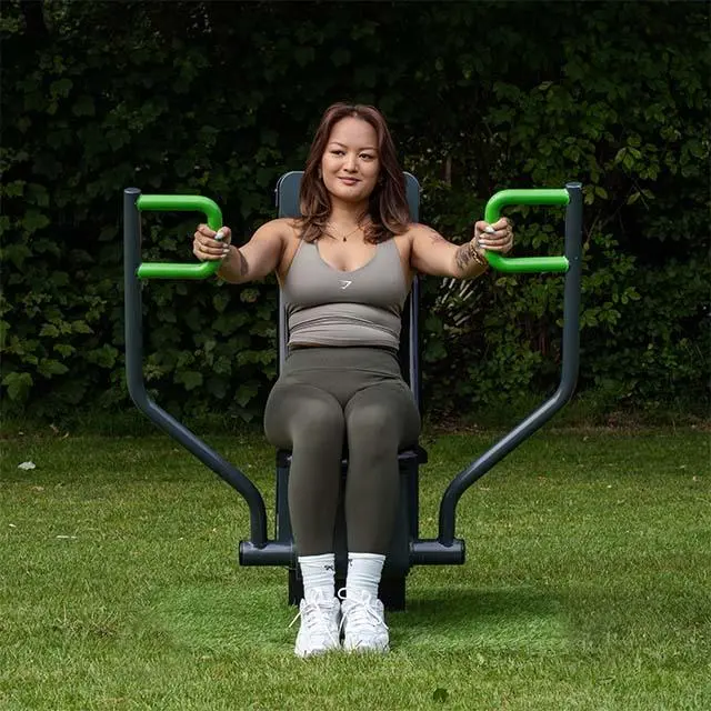 A woman in athletic wear uses an outdoor exercise machine in a grassy park, surrounded by trees. She looks focused and content.